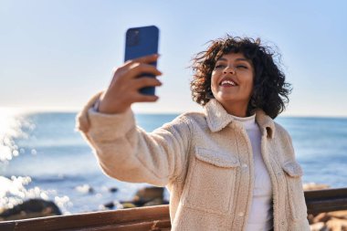 Young beautiful hispanic woman smiling confident make selfie by the smartphone at seaside