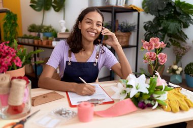 Young african american woman florist talking on smartphone writing on document at flower shop