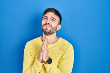 Hispanic man standing over blue background begging and praying with hands together with hope expression on face very emotional and worried. begging. 