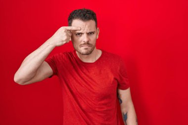 Young hispanic man standing over red background pointing unhappy to pimple on forehead, ugly infection of blackhead. acne and skin problem 