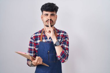Young hispanic man with beard wearing waiter apron holding clipboard asking to be quiet with finger on lips. silence and secret concept. 