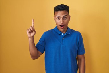 Young hispanic man standing over yellow background pointing finger up with successful idea. exited and happy. number one. 