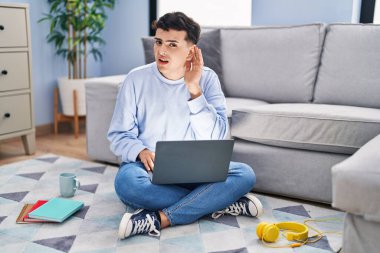 Non binary person studying using computer laptop sitting on the floor smiling with hand over ear listening an hearing to rumor or gossip. deafness concept. 