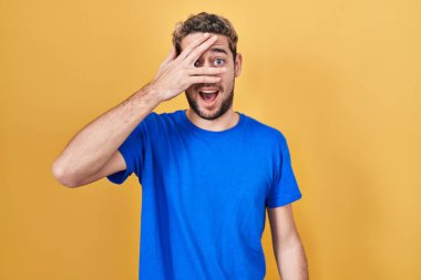 Hispanic man with beard standing over yellow background peeking in shock covering face and eyes with hand, looking through fingers with embarrassed expression. 