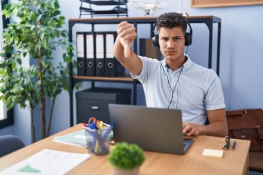 Young hispanic man working at the office wearing headphones looking unhappy and angry showing rejection and negative with thumbs down gesture. bad expression. 
