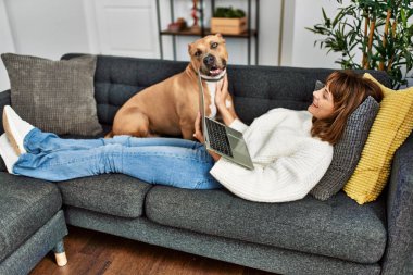 Genç beyaz kadın evdeki köpekle birlikte kanepede dizüstü bilgisayar kullanıyor.