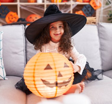 Adorable hispanic girl wearing halloween costume holding pumpkin basket lamp at home