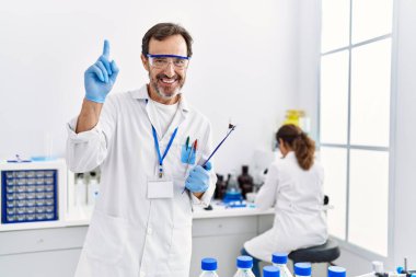 Middle age man working at scientist laboratory surprised with an idea or question pointing finger with happy face, number one 