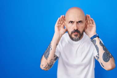 Mavi arka planda dikilen İspanyol dövmeli adam iki elini de kulak hareketi yaparken duymaya çalışıyor. Dedikoduya meraklı. İşitme sorunu, sağır 
