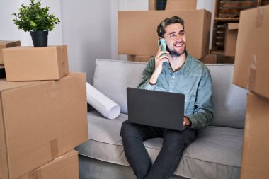 Young man using laptop talking on smartphone at new home