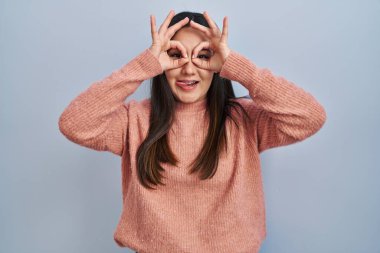 Young latin woman standing over blue background doing ok gesture like binoculars sticking tongue out, eyes looking through fingers. crazy expression. 