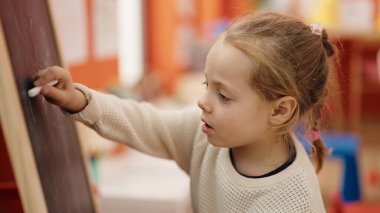 Adorable blonde girl preschool student drawing on blackboard at kindergarten