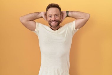 Middle age man with beard standing over yellow background relaxing and stretching, arms and hands behind head and neck smiling happy 