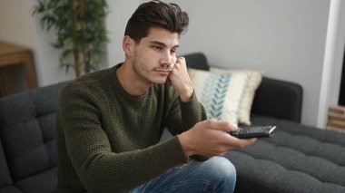 Young hispanic man watching tv sitting on sofa with boring expression at home