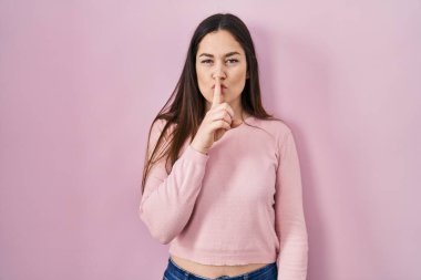 Genç esmer kadın pembe arka planda dikiliyor ve parmakları dudaklarında sessiz olmak istiyor. sessizlik ve gizli konsept. 