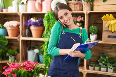 Young beautiful hispanic woman florist talking on smartphone writing on document at flower shop