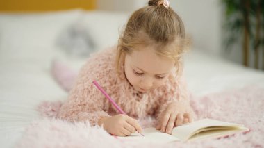Adorable blonde girl drawing on notebook lying on bed at bedroom