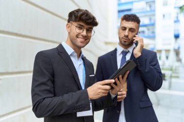 Two hispanic men business workers using touchpad talking on smartphone at street