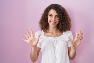 Hispanic woman with curly hair standing over pink background showing and pointing up with fingers number ten while smiling confident and happy. 