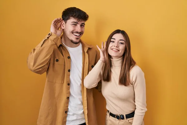 年轻的一对惊慌失措的夫妇站在黄色的背景上微笑着 两手空空地听着谣言或闲话 聋的概念 — 图库照片