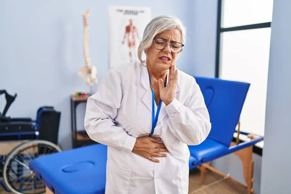 Middle age woman with grey hair working at pain recovery clinic with hand on stomach because indigestion, painful illness feeling unwell. ache concept.