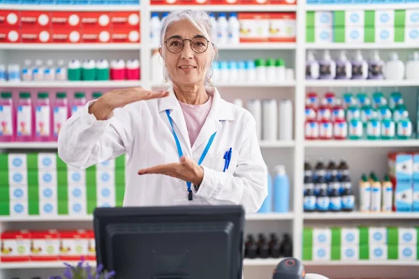 Middle age woman with tattoos working at pharmacy drugstore gesturing with hands showing big and large size sign, measure symbol. smiling looking at the camera. measuring concept.