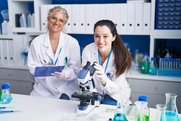 Two women scientists using microscope write on document at laboratory