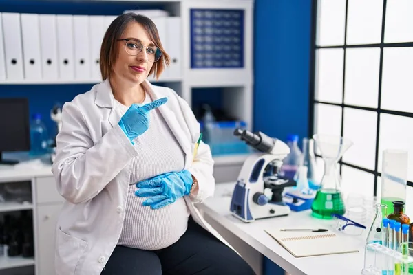Pregnant woman working at scientist laboratory pointing with hand finger to the side showing advertisement, serious and calm face