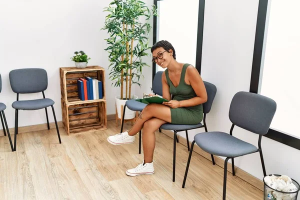Young hispanic woman writing on clipboard sitting on chair at waiting room
