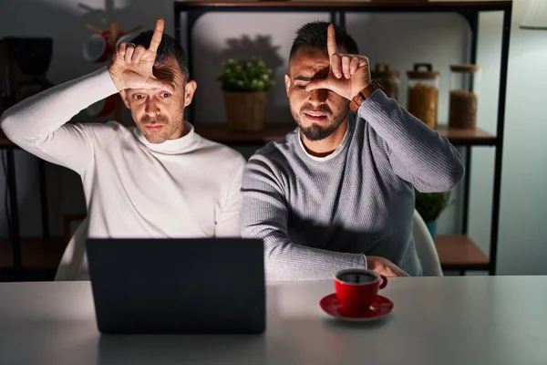 Homosexual couple using computer laptop making fun of people with fingers on forehead doing loser gesture mocking and insulting.