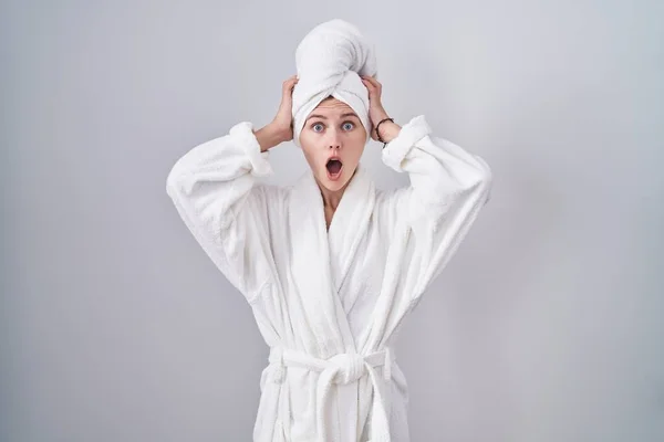Blonde caucasian woman wearing bathrobe crazy and scared with hands on head, afraid and surprised of shock with open mouth