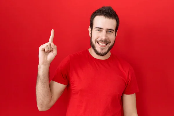 自信と幸せ笑顔ながらカジュアルな赤いTシャツを身に着けている若いヒスパニック系の男が現れ 指第1位で指摘します — ストック写真