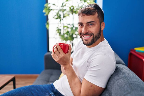 Young hispanic man drinking coffee sitting on sofa at home