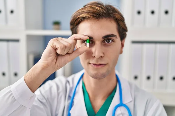 Young caucasian man doctor holding pill over eye at clinic