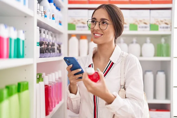薬局で薬瓶を持っているスマートフォンを使用して若い美しいヒスパニック系女性の顧客 — ストック写真