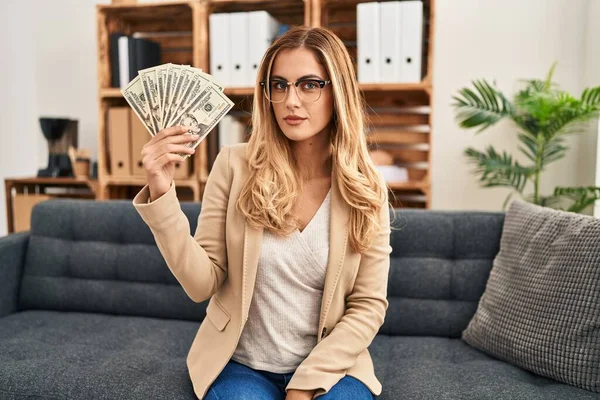 Young blonde therapist woman working at therapy office holding money thinking attitude and sober expression looking self confident 
