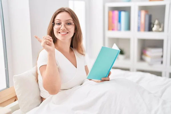 年轻的高加索女人坐在家里的床上看书 微笑着 手指手画脚地指向旁边 — 图库照片