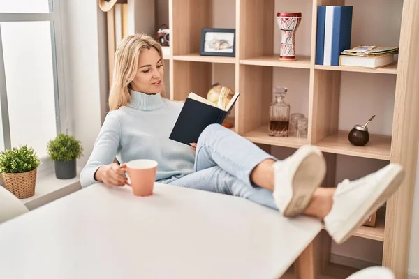Νεαρή Ξανθιά Γυναίκα Διαβάζει Βιβλίο Και Πίνει Καφέ Στο Σπίτι — Φωτογραφία Αρχείου