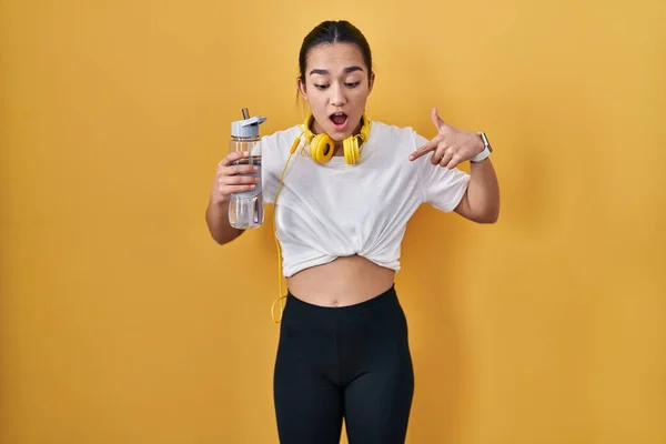 Young south asian woman wearing sportswear drinking water pointing down with fingers showing advertisement, surprised face and open mouth
