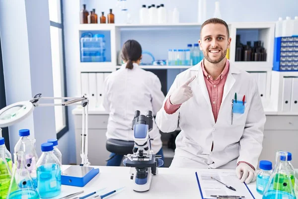 Young man working at scientist laboratory smiling happy pointing with hand and finger