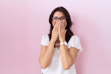 Middle age hispanic woman wearing casual white t shirt and glasses laughing and embarrassed giggle covering mouth with hands, gossip and scandal concept 