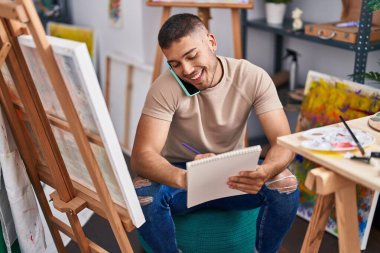 Young hispanic man artist talking on the smartphone drawing on notebook at art studio