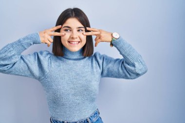 Mavi arka planda dikilen genç İspanyol kadın barış sembolü yapıyor. Yüzünde parmakları, gülümseyerek zaferi gösteriyor. 