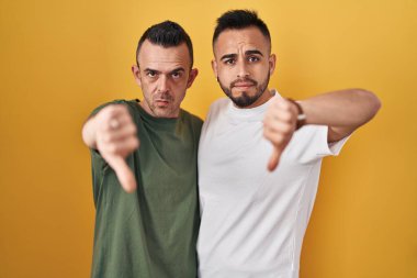 Homosexual couple standing over yellow background looking unhappy and angry showing rejection and negative with thumbs down gesture. bad expression. 