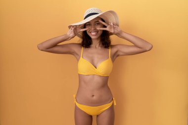 Young hispanic woman wearing bikini and summer hat doing peace symbol with fingers over face, smiling cheerful showing victory 