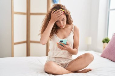 Genç, güzel, İspanyol bir kadın yatak odasında endişeli bir ifadeyle akıllı telefon kullanıyor.