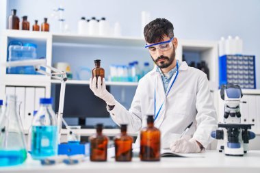 Young hispanic man scientist writing on notebook holding bottle at laboratory