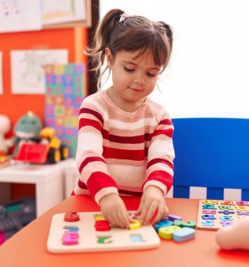 Adorable hispanic girl playing with maths puzzle game standing at kindergarten