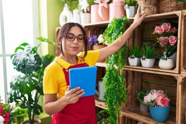 Young beautiful arab woman florist using touchpad holding plant of shelving at flower shop