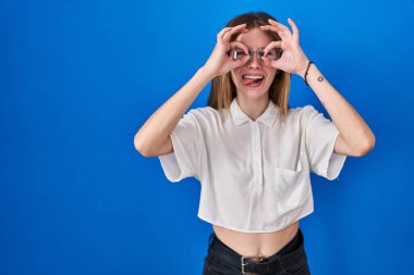 Beautiful woman standing over blue background doing ok gesture like binoculars sticking tongue out, eyes looking through fingers. crazy expression. 